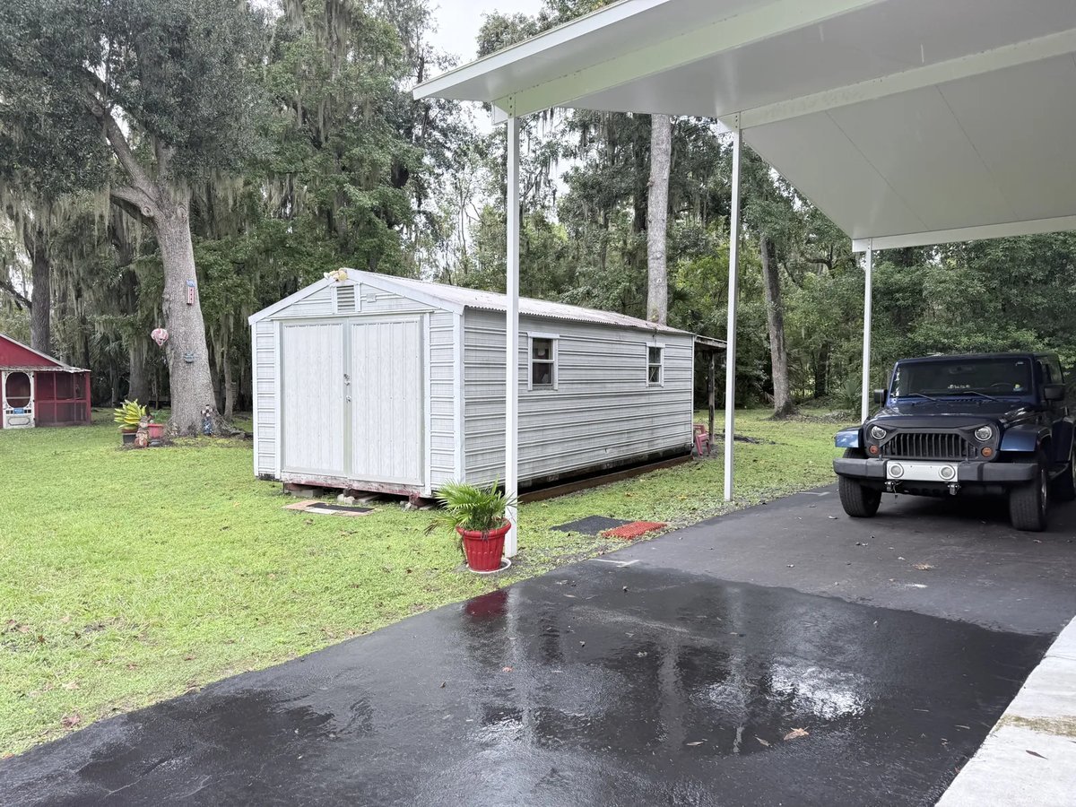 Florida yard with a storage shed, mature oaks hung with Spanish moss, and a carport on a wet driveway